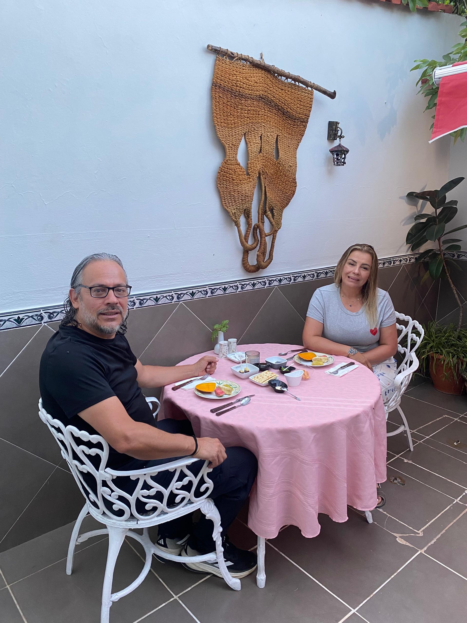 A couple at a round pink-clothed table on the terrace, woven art hanging behind them